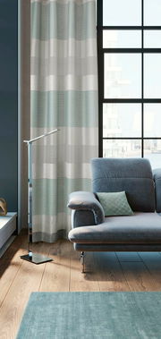 Moderner Ösenvorhang in Grau- und Grüntönen vor einem großen Fenster, neben einem grauen Sofa in einem stilvollen Wohnzimmer. Perspektive von der Seite.