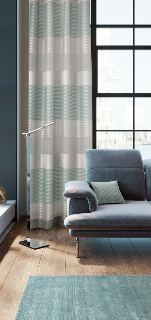 Moderner Ösenvorhang in Grau- und Grüntönen vor einem großen Fenster, neben einem grauen Sofa in einem stilvollen Wohnzimmer. Perspektive von der Seite.