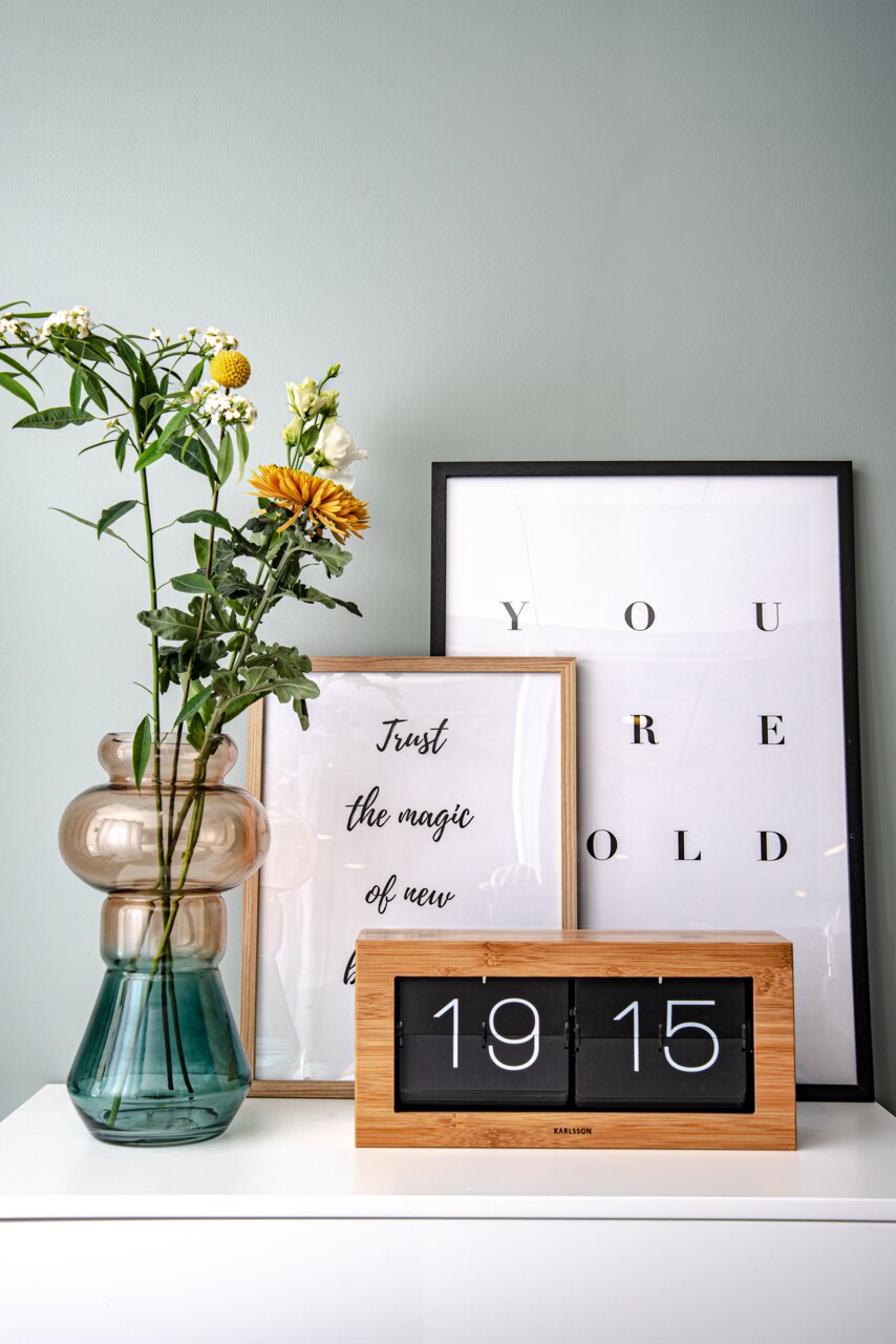 Een bamboe-klok op een witte plank, omringd door een vaas met bloemen en ingelijste posters, gezien vanuit een vooraanzicht.