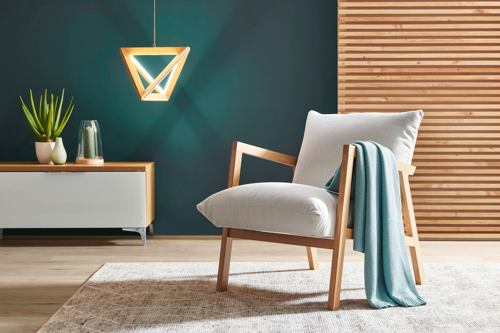 Moderner Sessel mit Holzrahmen und hellgrauem Polster, dekoriert mit einer türkisfarbenen Decke, in einem stilvollen Wohnzimmer mit hölzernem Wandpaneel und hängender Lampe, seitliche Perspektive.