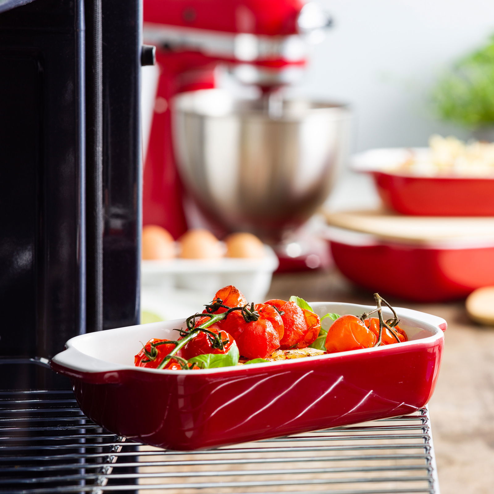 Rote Auflaufform mit Deckel, gefüllt mit Tomaten und Basilikum, in einem Ofen. Seitliche Perspektive.