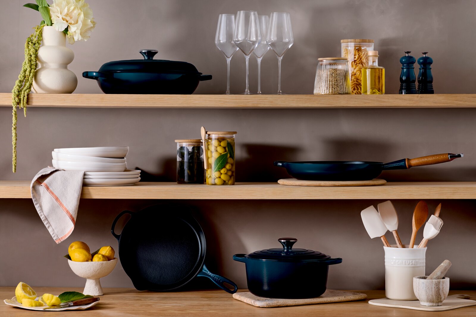 LE CREUSET Koekenpan 28 cm SIGNATURE Sfeervolle keukenplank met donkerblauw gietijzeren kookgerei (braadpan met deksel, koekenpan en grillpan), wijnglazen, voorraadpotten en keukengerei op houten planken tegen een beige muur, gefotografeerd in frontaal aanzicht op ooghoogte.