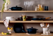 Sfeervolle keukenplank met donkerblauw gietijzeren kookgerei (braadpan met deksel, koekenpan en grillpan), wijnglazen, voorraadpotten en keukengerei op houten planken tegen een beige muur, gefotografeerd in frontaal aanzicht op ooghoogte.