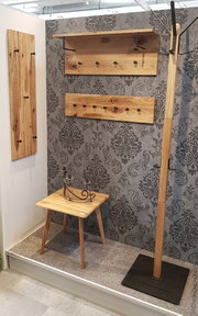 Ausstellungsansicht in schräger Frontperspektive: rustikale Wandgarderobe aus hellem Massivholz mit schwarzen Metallhaken und Ablage, daneben schlanke Standgarderobe auf schwarzem Sockel sowie kleiner Holztisch vor gemusterter Tapete.