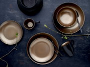 Modernes Geschirrset in Bronze- und Schwarztönen mit Tellern, Schalen und Tassen auf dunklem Untergrund, Draufsicht (Flatlay)