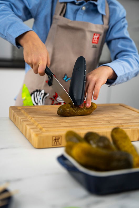 Kind schneidet mit einem blauen Kinderkochmesser eine Gurke auf einem Holzbrett, Vorderansicht