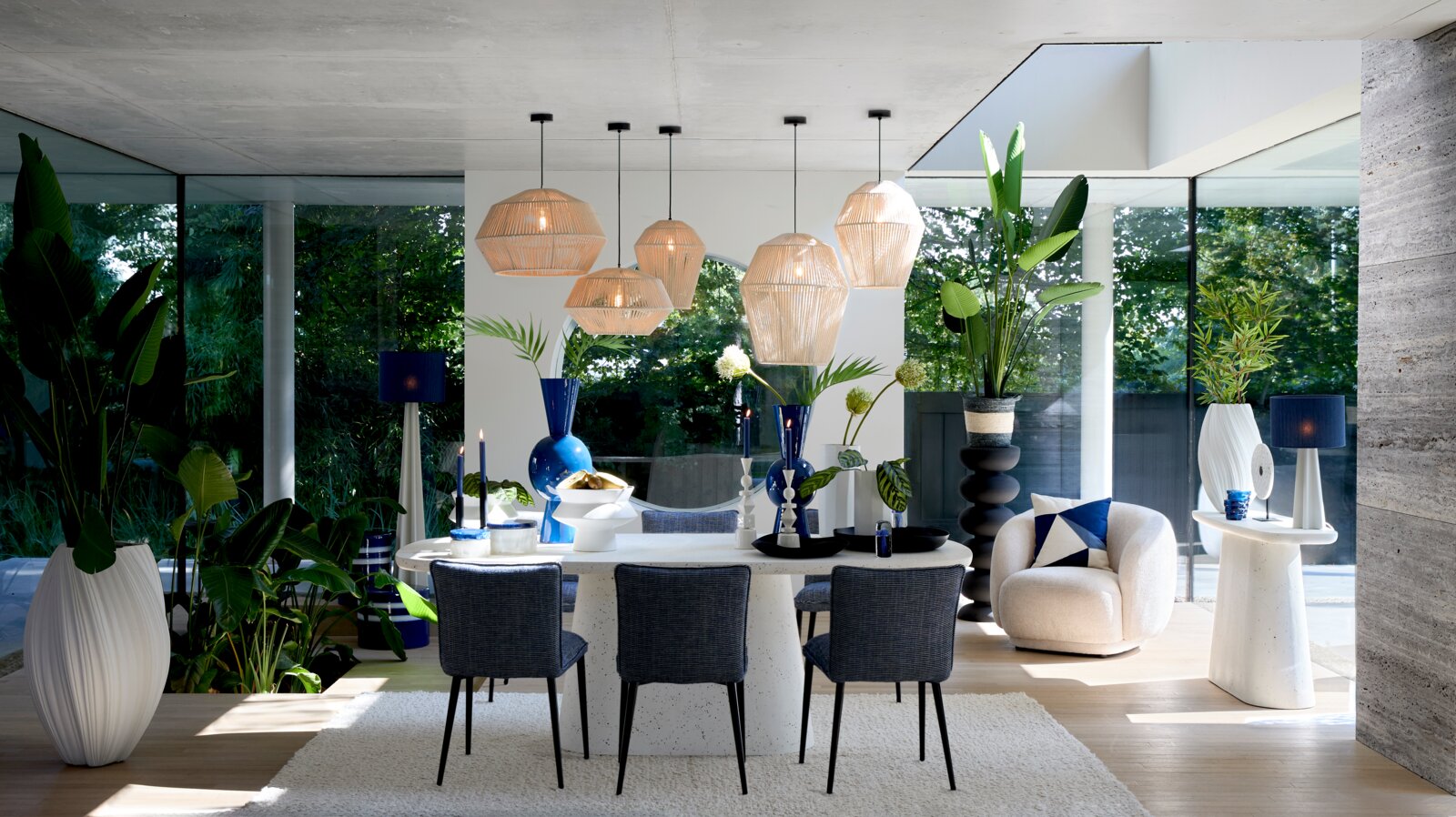 Frontaal perspectief op een luxe, modern interieur met witte eettafel, donkerblauwe stoelen, rotan hanglampen, een crèmekleurige fauteuil en weelderige groene planten.