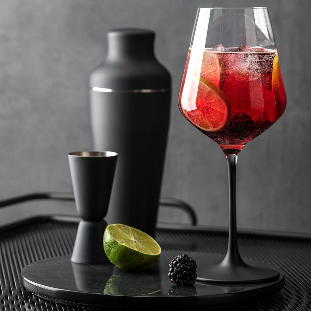Ein eleganter Rotweinkelch gefüllt mit einem roten Getränk, garniert mit Limettenscheiben und Beeren, im Vordergrund. Im Hintergrund sind ein schwarzer Cocktailshaker und ein Messbecher zu sehen. Perspektive: seitlich.