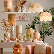 Ein stilvoll eingerichteter Raum mit verschiedenen Lampenschirmen aus natürlichen Materialien, darunter hängende und stehende Modelle, umgeben von dekorativen Vasen und Pflanzen. Die Perspektive zeigt eine frontale Ansicht auf die Tischdekoration und die 