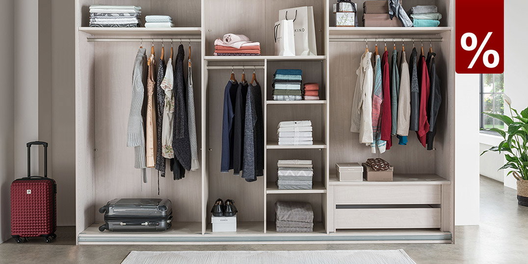 Großer offener Kleiderschrank mit Fächern, Kleiderstangen und Schubladen, ordentlich gefüllt mit Kleidung, Kissen und Boxen, daneben ein Koffer