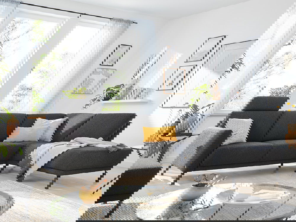 Das Bild zeigt ein gemütliches Wohnzimmer mit einem grauen Ecksofa. Auf dem Sofa liegen bunte Kissen und eine Decke. Im Hintergrund gibt es helle Fenster mit weißen Vorhängen. An der Wand hängen Bilder und eine kleine Regalablage mit Dekoration.