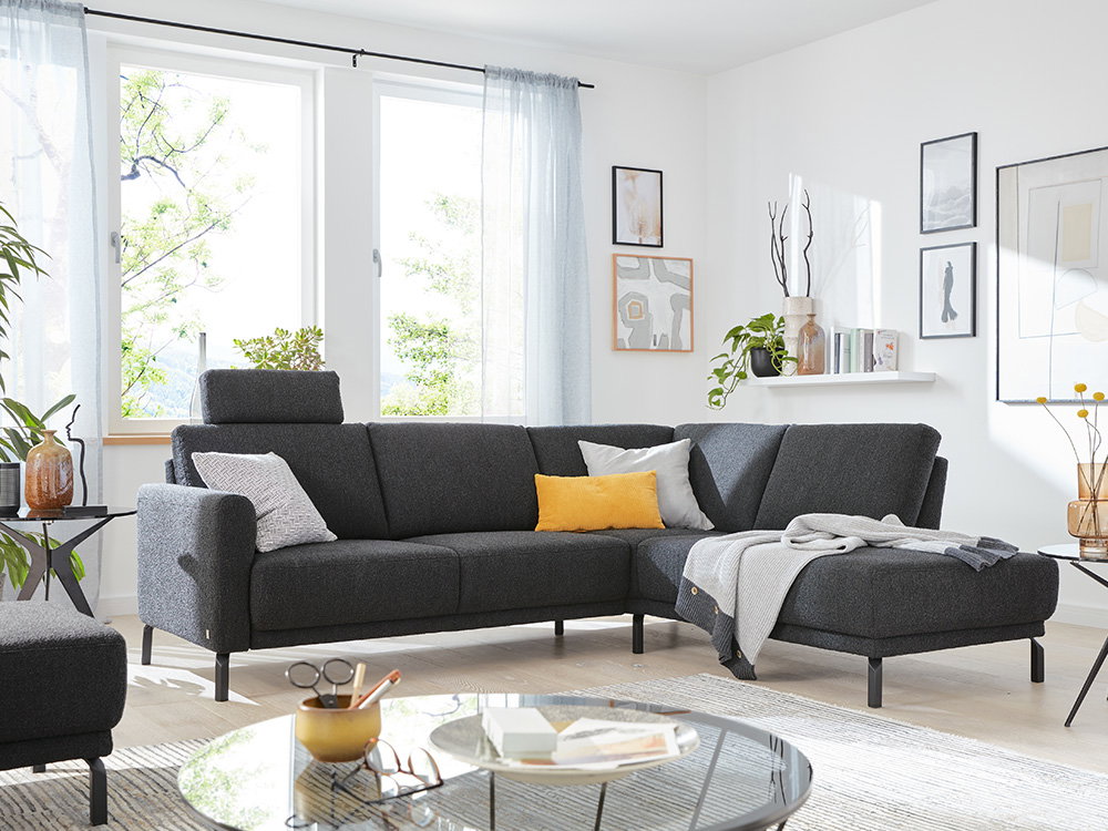 Das Bild zeigt ein gemütliches Wohnzimmer mit einem grauen Ecksofa. Auf dem Sofa liegen bunte Kissen und eine Decke. Im Hintergrund gibt es helle Fenster mit weißen Vorhängen. An der Wand hängen Bilder und eine kleine Regalablage mit Dekoration.