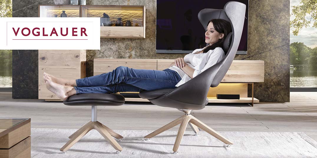 Eine Frau entspannt in einem modernen Sessel mit Fußhocker in einem stilvollen Wohnzimmer mit Holzmöbeln.
