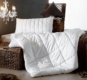 Weiße Faserdecke Duo ANTIGUA von billerbeck, drapiert auf einem Rattankorb, mit passendem Kissen, seitliche Perspektive.