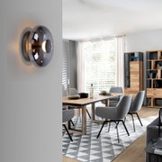 Moderne runde Wandleuchte aus Rauchglas mit warmweißem Licht an hellgrauer Wand, seitliche Nahaufnahme in einem hellen Esszimmer-Interieur