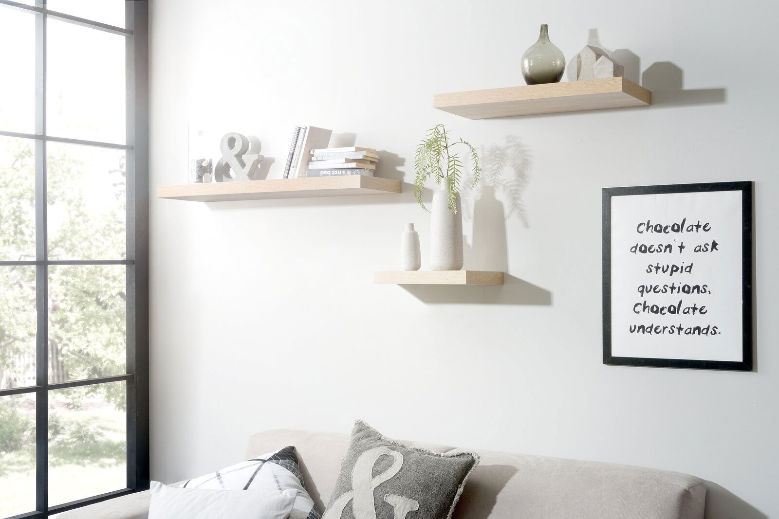 Drei schwebende Wandregale in hellem Holzton, dekoriert mit Büchern, Vasen und Dekorationsobjekten, in einem modernen Wohnzimmer mit Sofa und großem Fenster, seitliche Perspektive.