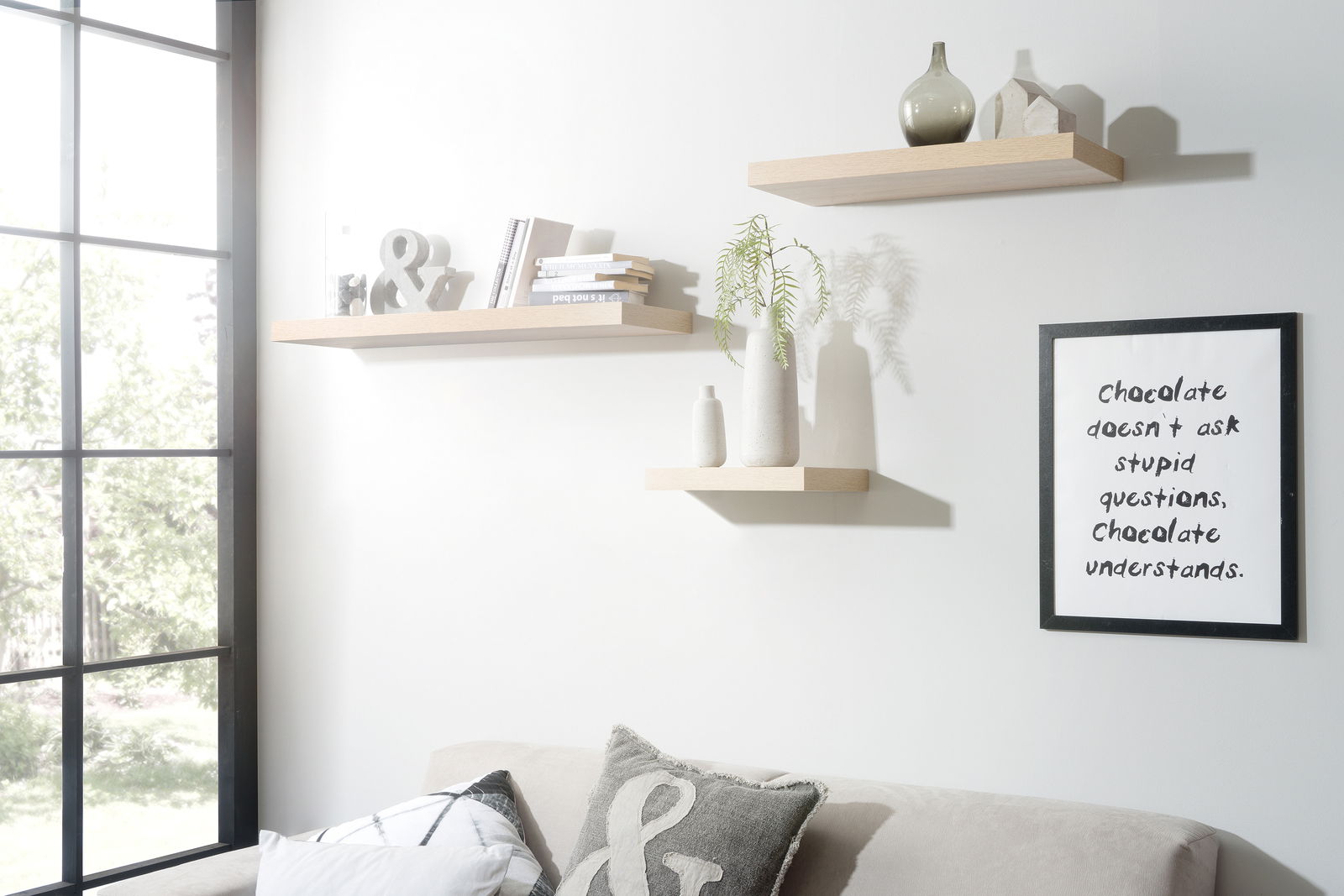 Drei schwebende Wandregale in hellem Holzton, dekoriert mit Büchern, Vasen und Dekorationsobjekten, in einem modernen Wohnzimmer mit Sofa und großem Fenster, seitliche Perspektive.