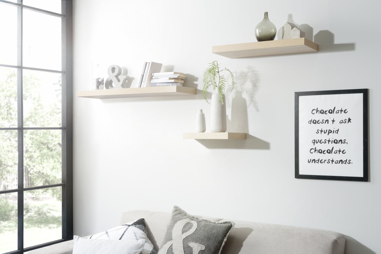 Drei schwebende Wandregale in hellem Holzton, dekoriert mit Büchern, Vasen und Dekorationsobjekten, in einem modernen Wohnzimmer mit Sofa und großem Fenster, seitliche Perspektive.