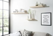Drei schwebende Wandregale in hellem Holzton, dekoriert mit Büchern, Vasen und Dekorationsobjekten, in einem modernen Wohnzimmer mit Sofa und großem Fenster, seitliche Perspektive.