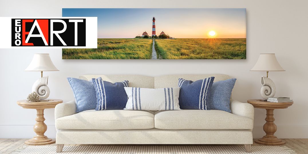 Ein gemütliches Wohnzimmer mit einem weißen Sofa und blauen Kissen, auf dem ein großes Kunstwerk mit einem Leuchtturm bei Sonnenuntergang zu sehen ist.