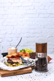 Pfeffermühle AKASIA aus Holz und Glas, gefüllt mit Pfefferkörnern, auf einem Tisch neben einem Burger, seitliche Perspektive.