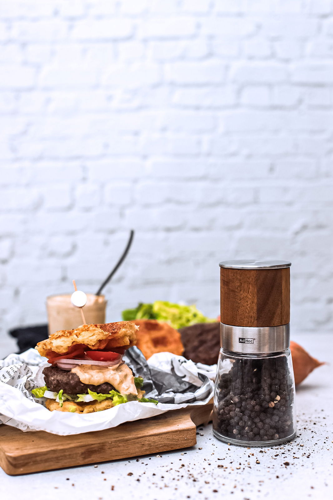 Pfeffermühle AKASIA aus Holz und Glas, gefüllt mit Pfefferkörnern, auf einem Tisch neben einem Burger, seitliche Perspektive.
