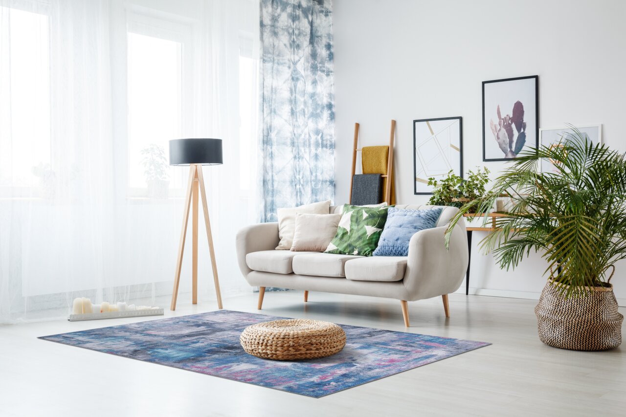 Zurbrüggen Tapijt PRIMA Moderne woonkamer met op de voorgrond een blauw en paars tapijt, een beige bank met kleurrijke kussens, een staande lamp en planten op de achtergrond. Aanzicht van voren.
