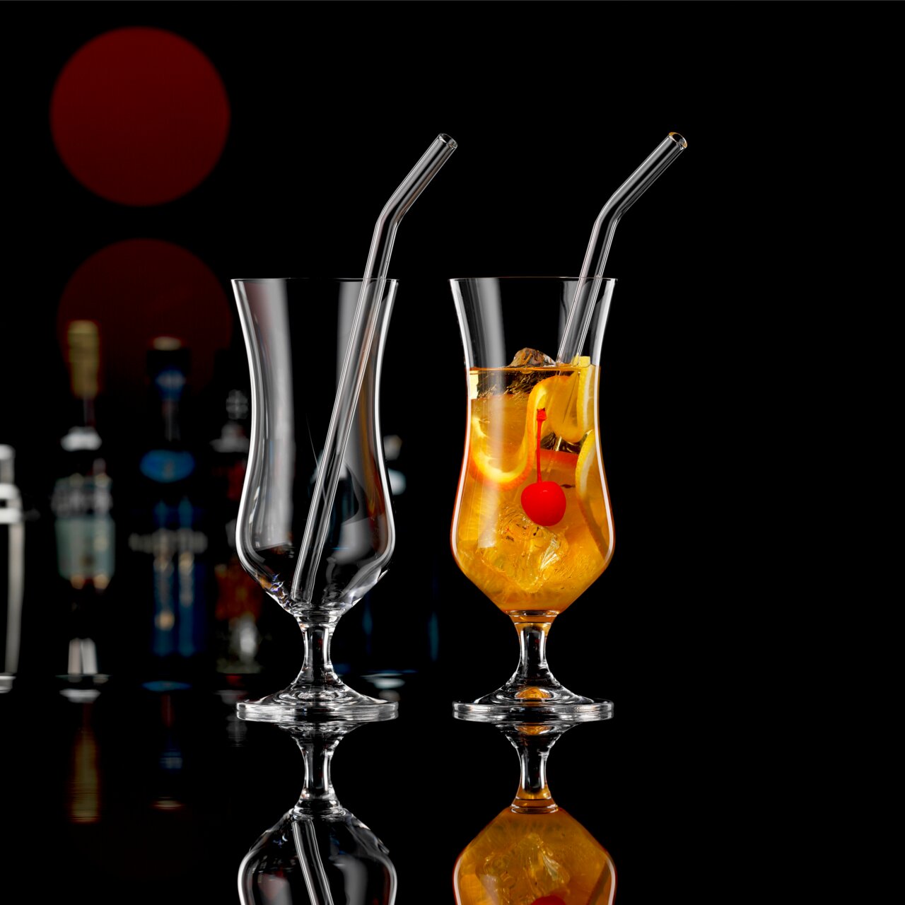 Novel Cocktailgläser 2er-Set mit Trinkhalm Zwei elegante Cocktailgläser mit Trinkhalm, eines leer und eines gefüllt mit einem Getränk und einer Kirsche, vor einem dunklen Hintergrund mit Flaschen im Hintergrund, seitliche Perspektive.