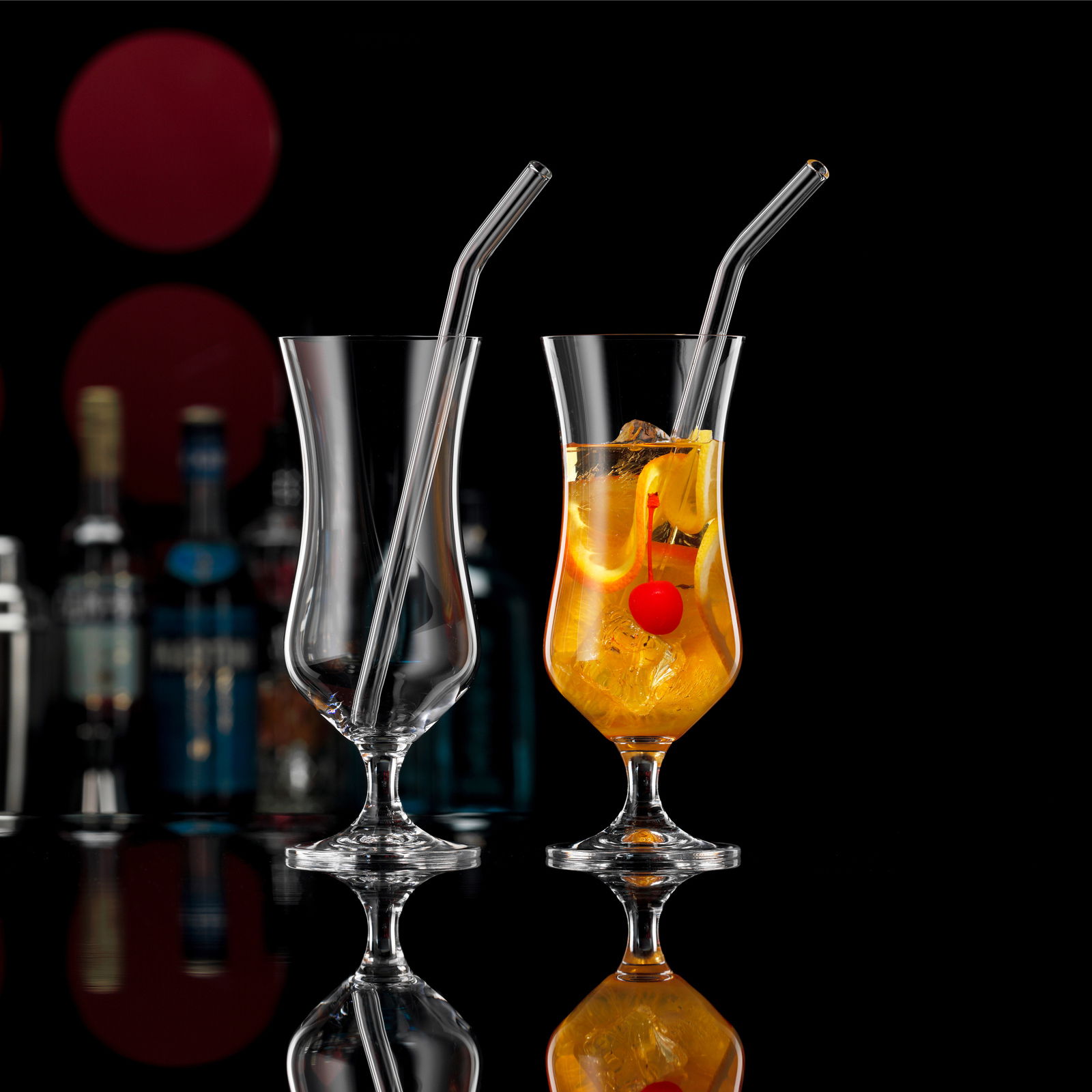 Zwei elegante Cocktailgläser mit Trinkhalm, eines leer und eines gefüllt mit einem Getränk und einer Kirsche, vor einem dunklen Hintergrund mit Flaschen im Hintergrund, seitliche Perspektive.