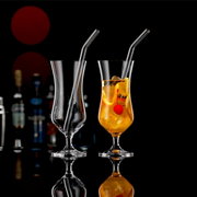 Twee elegante cocktailglazen met rietje, één leeg en één gevuld met een drankje en een kers, tegen een donkere achtergrond met flessen op de achtergrond, zijaanzicht.