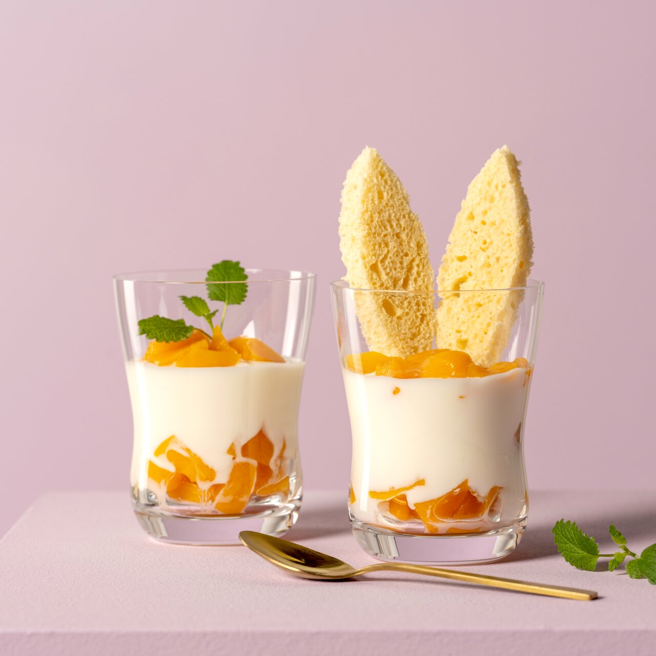 Zwei durchsichtige Becher mit 360ml Fassungsvermögen, gefüllt mit Joghurt und Pfirsichstücken, auf einem rosa Hintergrund. Vorderansicht.