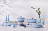 Eine Kollektion von hellblauen Küchenutensilien, darunter Bräter, Pfannen und Gewürzmühlen, auf einer hellen Oberfläche mit einer dekorativen Vase im Hintergrund. Frontalansicht.