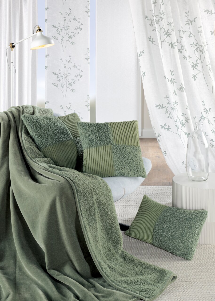 Gemütliche Wohnzimmerszene mit grünen Kissen und Decke auf einem Sofa, im Hintergrund weiße Vorhänge mit floralem Muster, seitliche Perspektive.