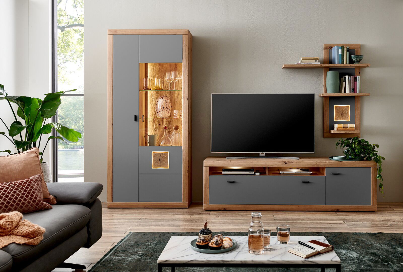 Moderne Wohnzimmer-Wandkombination mit grauen und holzfarbenen Elementen, bestehend aus einem hohen Schrank mit Glaseinsatz, einem TV-Board und einem Wandregal. Perspektive: Frontalansicht.