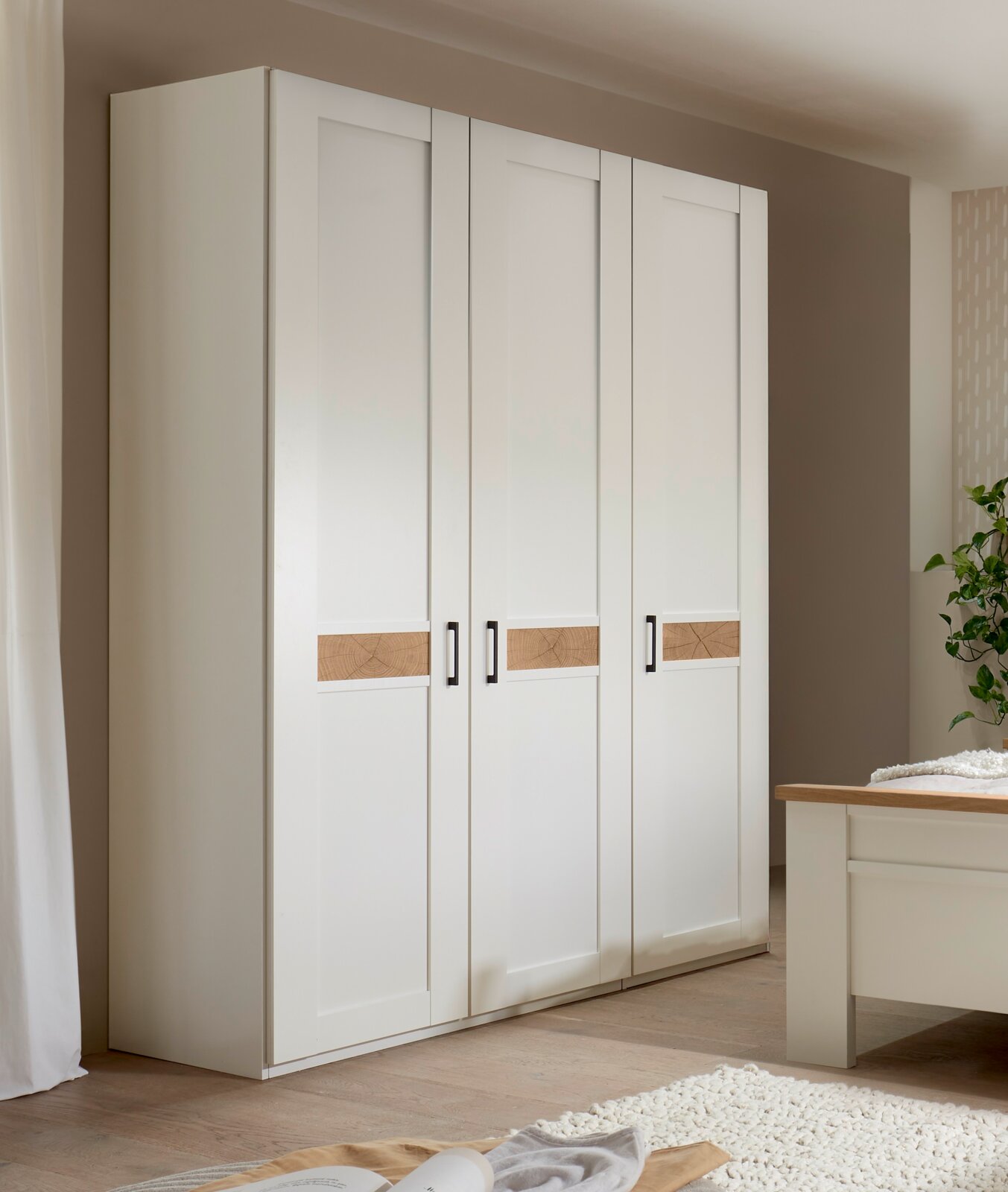Weißer 4-türiger Kleiderschrank im Landhausstil mit horizontalen Holz-Akzentstreifen und schwarzen Griffen, fotografiert in leichter Schrägansicht von vorn in einem Schlafzimmer.