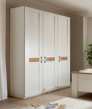 Weißer 4-türiger Kleiderschrank im Landhausstil mit horizontalen Holz-Akzentstreifen und schwarzen Griffen, fotografiert in leichter Schrägansicht von vorn in einem Schlafzimmer.