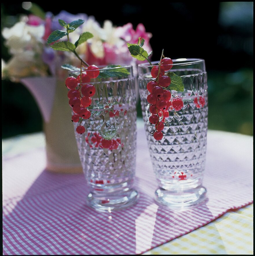 Zwei Longdrinkgläser aus der Boston-Serie mit facettiertem Design, gefüllt mit Wasser und dekoriert mit roten Johannisbeeren und Minzblättern, auf einem Tisch mit gestreifter Tischdecke, seitliche Perspektive.
