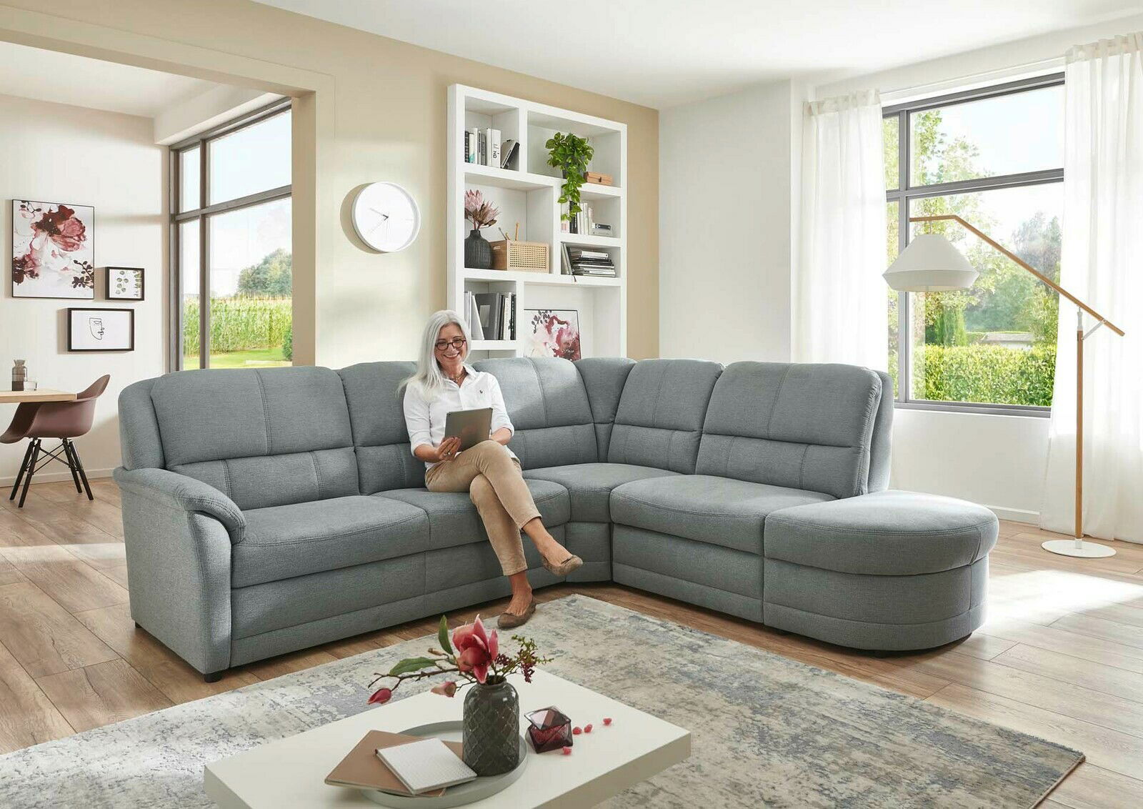 Gemütliche Polsterrundecke in einem modernen Wohnzimmer, seitliche Perspektive. Eine Person sitzt entspannt auf dem Sofa mit einem Tablet in der Hand.