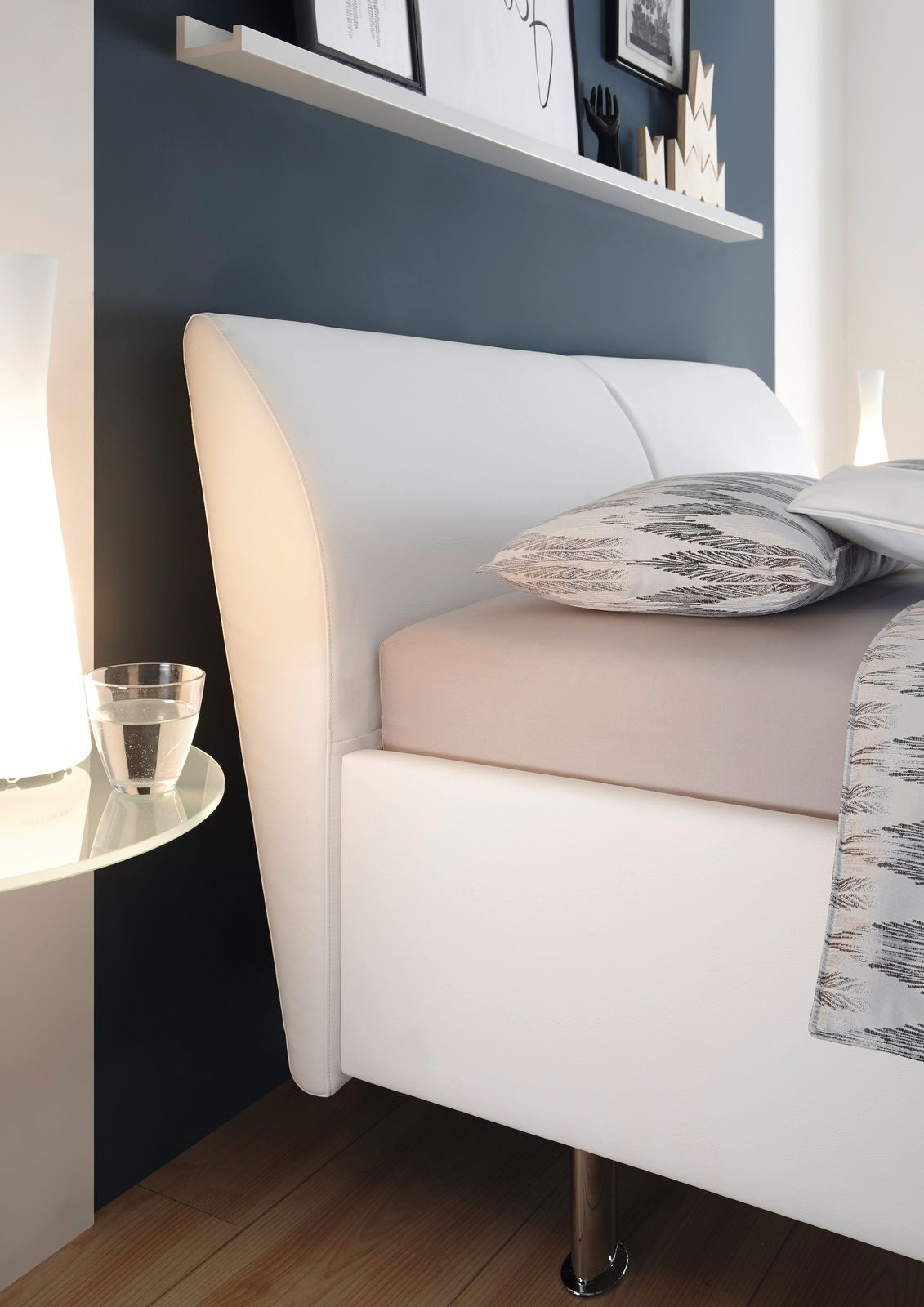 Seitliche Ansicht eines modernen Polsterbettgestells mit weißem Kopfteil und Bettgestell, dekoriert mit gemusterten Kissen und Decke, neben einem Nachttisch mit Glas und Lampe.