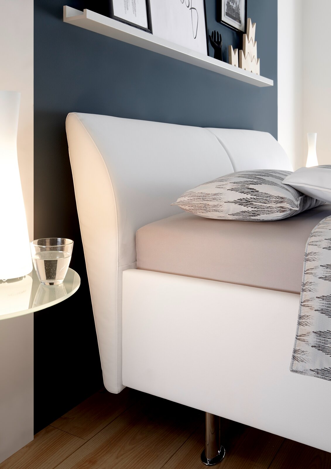 Seitliche Ansicht eines modernen Polsterbettgestells mit weißem Kopfteil und Bettgestell, dekoriert mit gemusterten Kissen und Decke, neben einem Nachttisch mit Glas und Lampe.