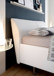 Zijaanzicht van een modern gestoffeerd bedframe met een wit hoofdeinde en bedframe, versierd met kussens en een deken met patroon, naast een nachtkastje met glazen blad en een lamp.
