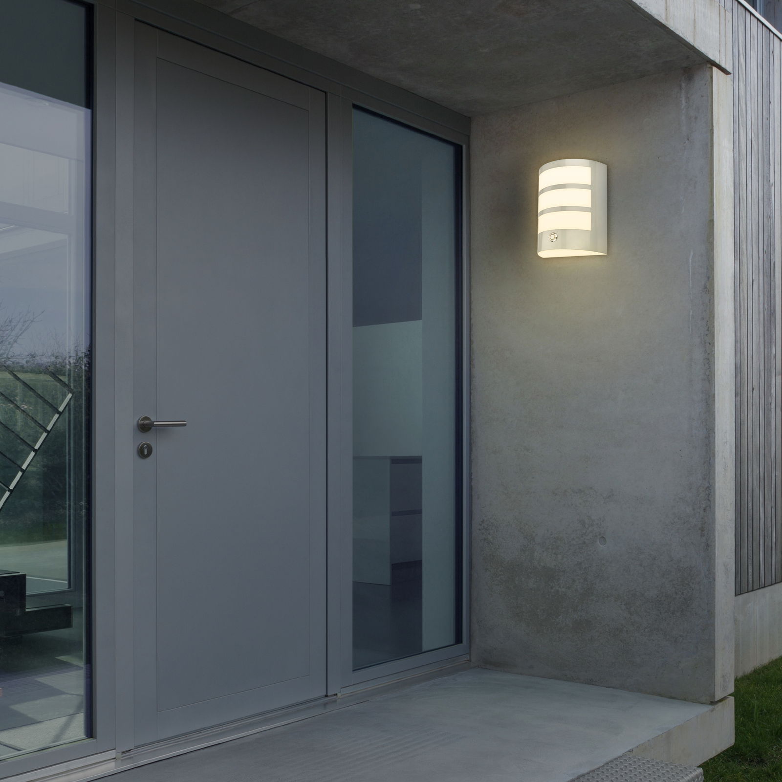Moderne Akku-Außenwandleuchte von Briloner, montiert an einer Hauswand neben einer Tür, in eingeschaltetem Zustand bei Dämmerung.
