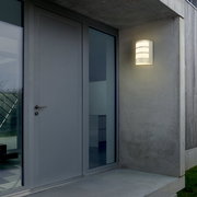 Moderne Akku-Außenwandleuchte von Briloner, montiert an einer Hauswand neben einer Tür, in eingeschaltetem Zustand bei Dämmerung.