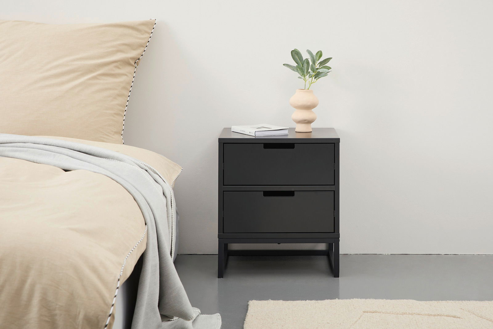 Schwarze Nachtkonsole mit zwei Schubladen, dekoriert mit einer Vase und einem Buch, seitliche Perspektive.