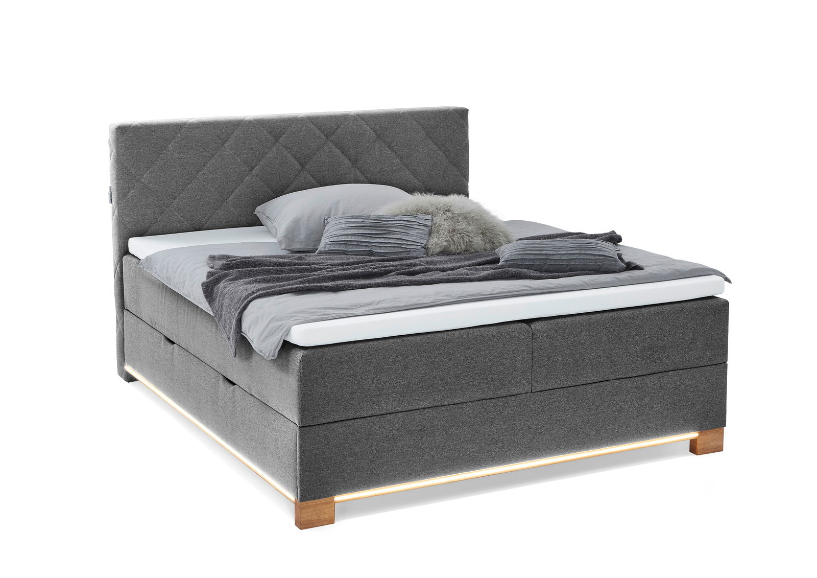 Graues Boxspringbett mit gestepptem Kopfteil, seitlicher Perspektive, dekoriert mit Kissen und Decken.