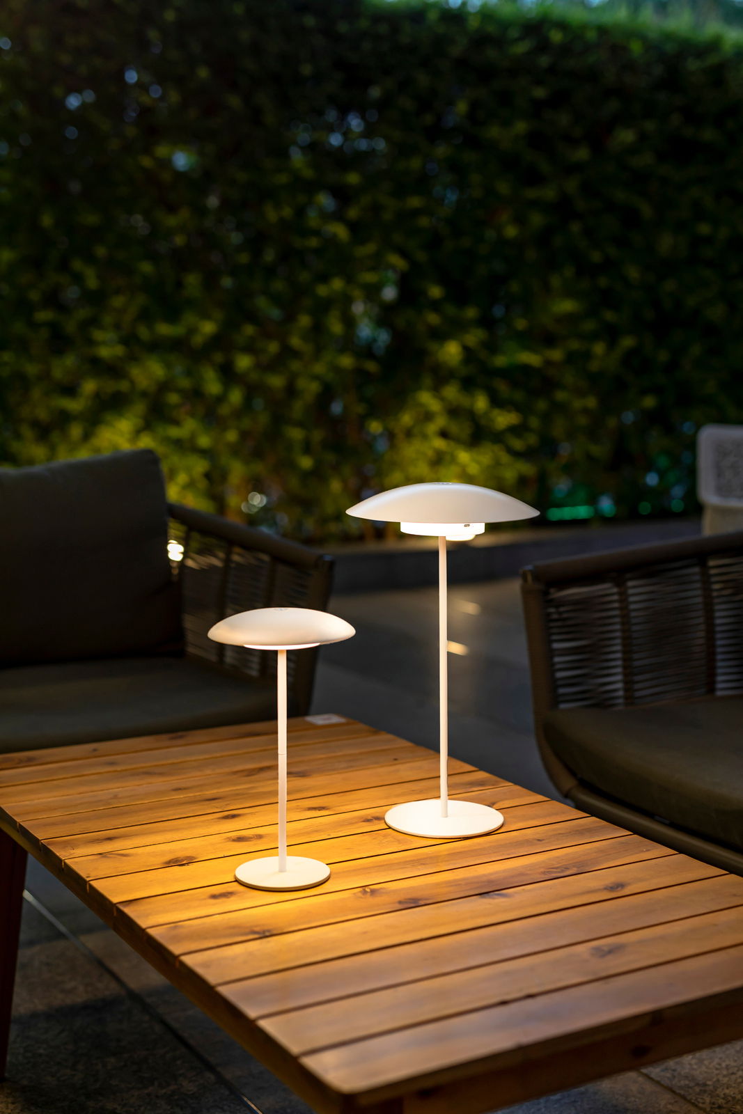 Zwei Akku-Tischleuchten SARDINIA von New Garden auf einem Holztisch im Freien, bei Dämmerung beleuchtet, mit Blick von schräg oben.