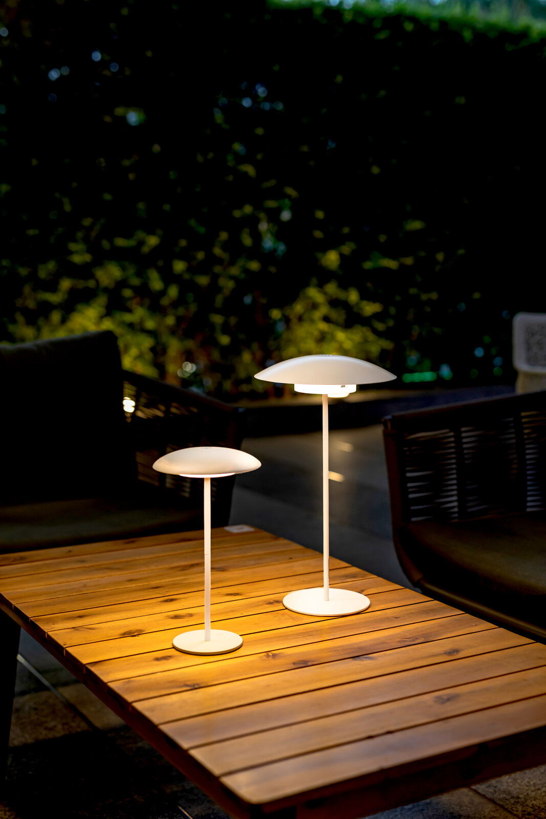 Zwei Akku-Tischleuchten SARDINIA von New Garden auf einem Holztisch im Freien, bei Dämmerung beleuchtet, mit Blick von schräg oben.