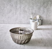 Gugelhupfform aus Metall mit einem Durchmesser von 24 cm, auf einer Küchenarbeitsplatte stehend, im Hintergrund Küchengeräte und ein Krug. Perspektive von vorne.