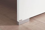 Nahaufnahme einer weißen Möbelkante mit grauem Sockel auf Holzboden, perspektivisch von der Seite aufgenommen.
