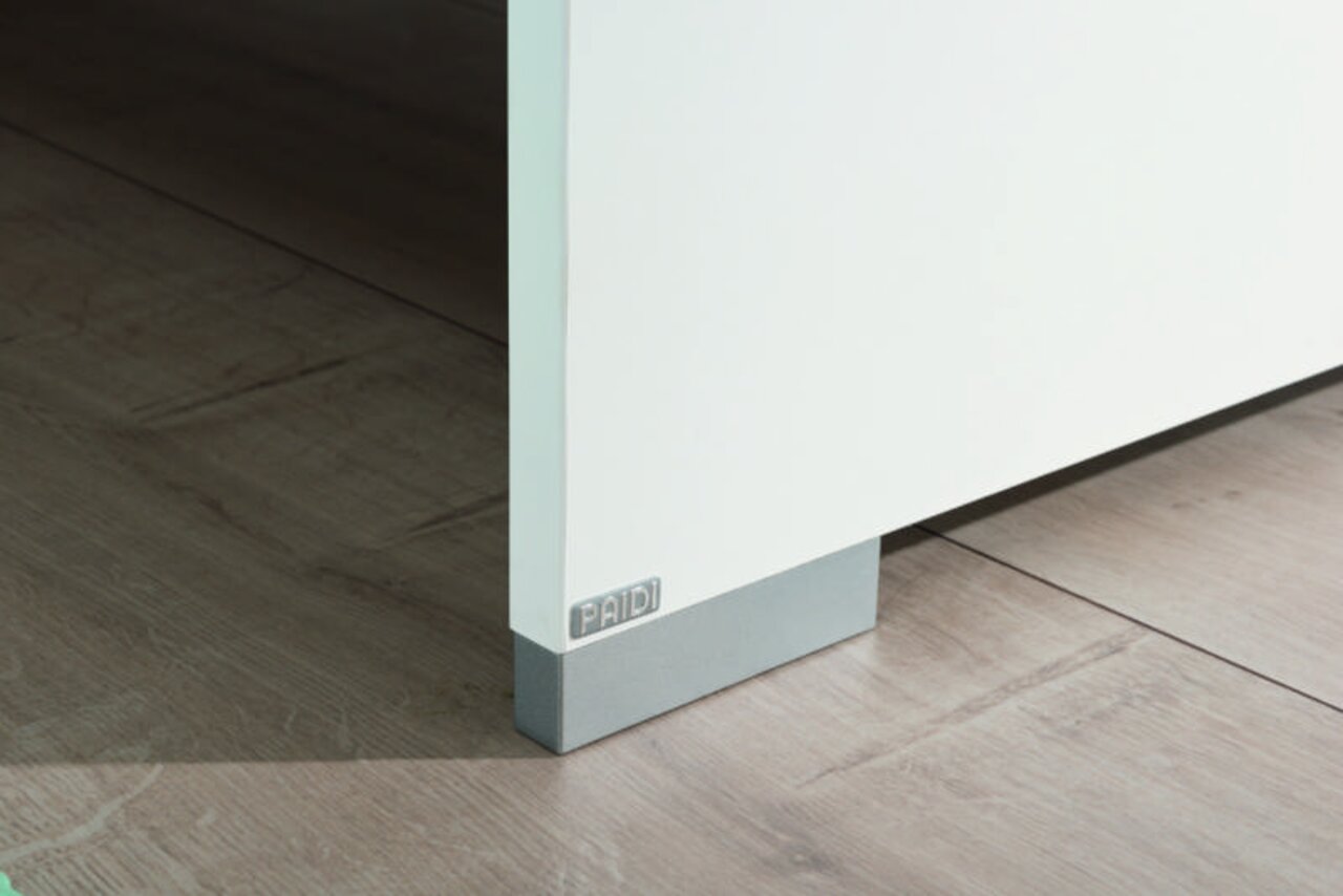 Nahaufnahme einer weißen Möbelkante mit grauem Sockel auf Holzboden, perspektivisch von der Seite aufgenommen.